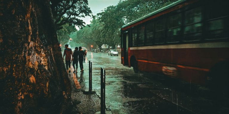 a group of people walking down a sidewalk next to a bus during mumbai rains